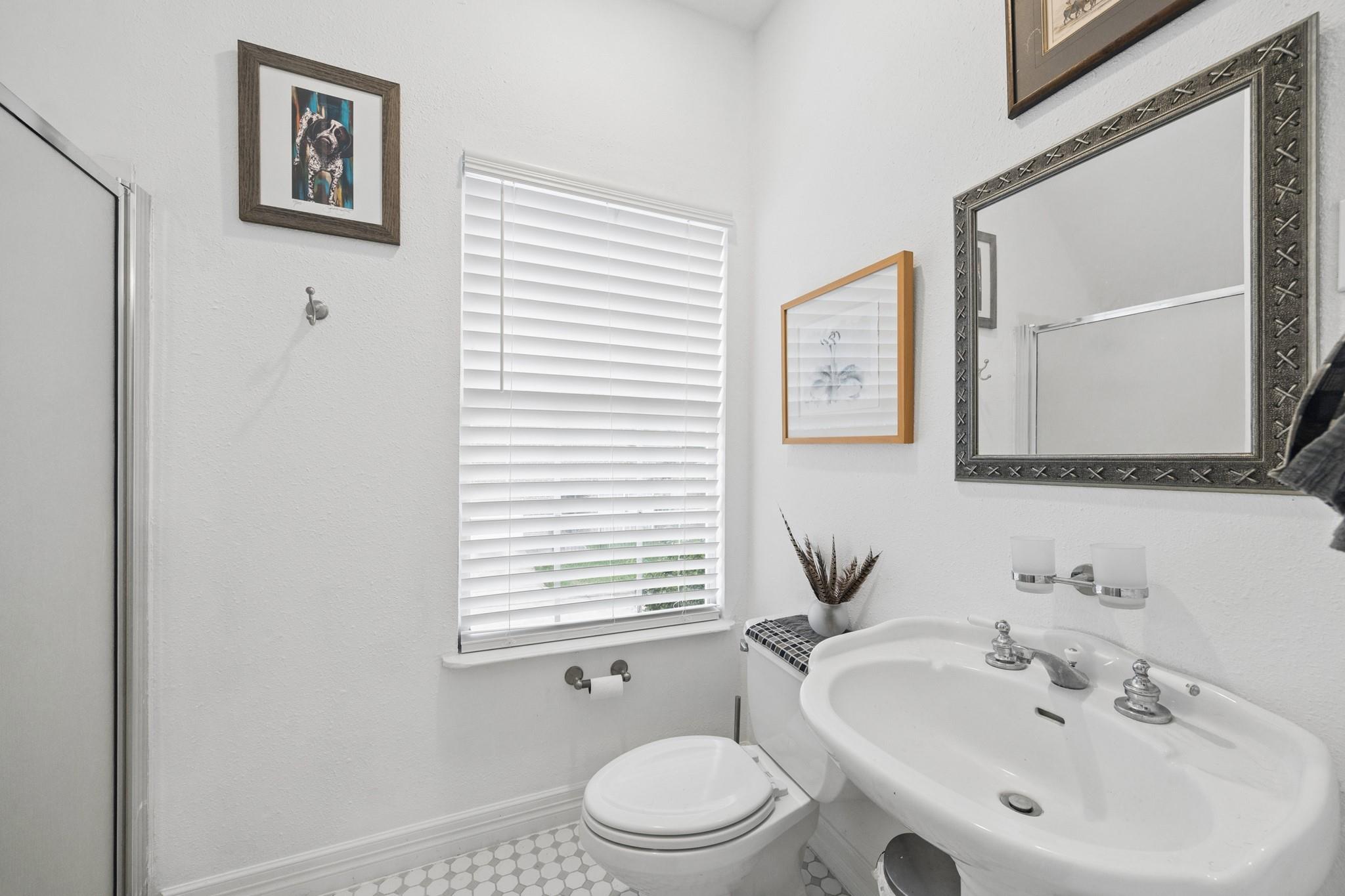 Bright bathroom with a pedestal sink, toilet, glass shower enclosure, and window with white blinds.