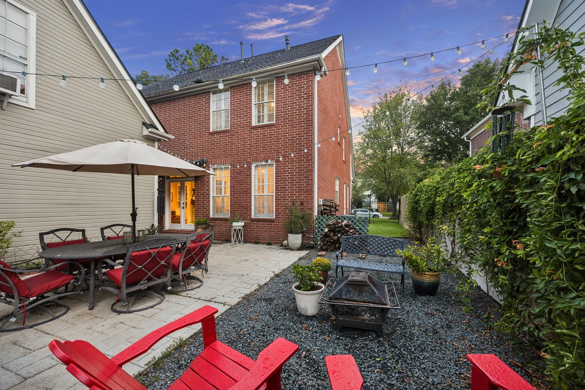 Backyard patio with red brick home, umbrella-covered dining table, string lights, and gravel seating area.