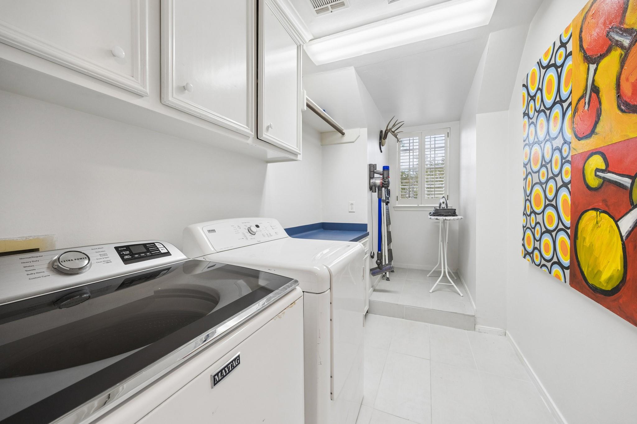 Laundry room with white Maytag washer and dryer, upper cabinets, shuttered window, and colorful wall art.