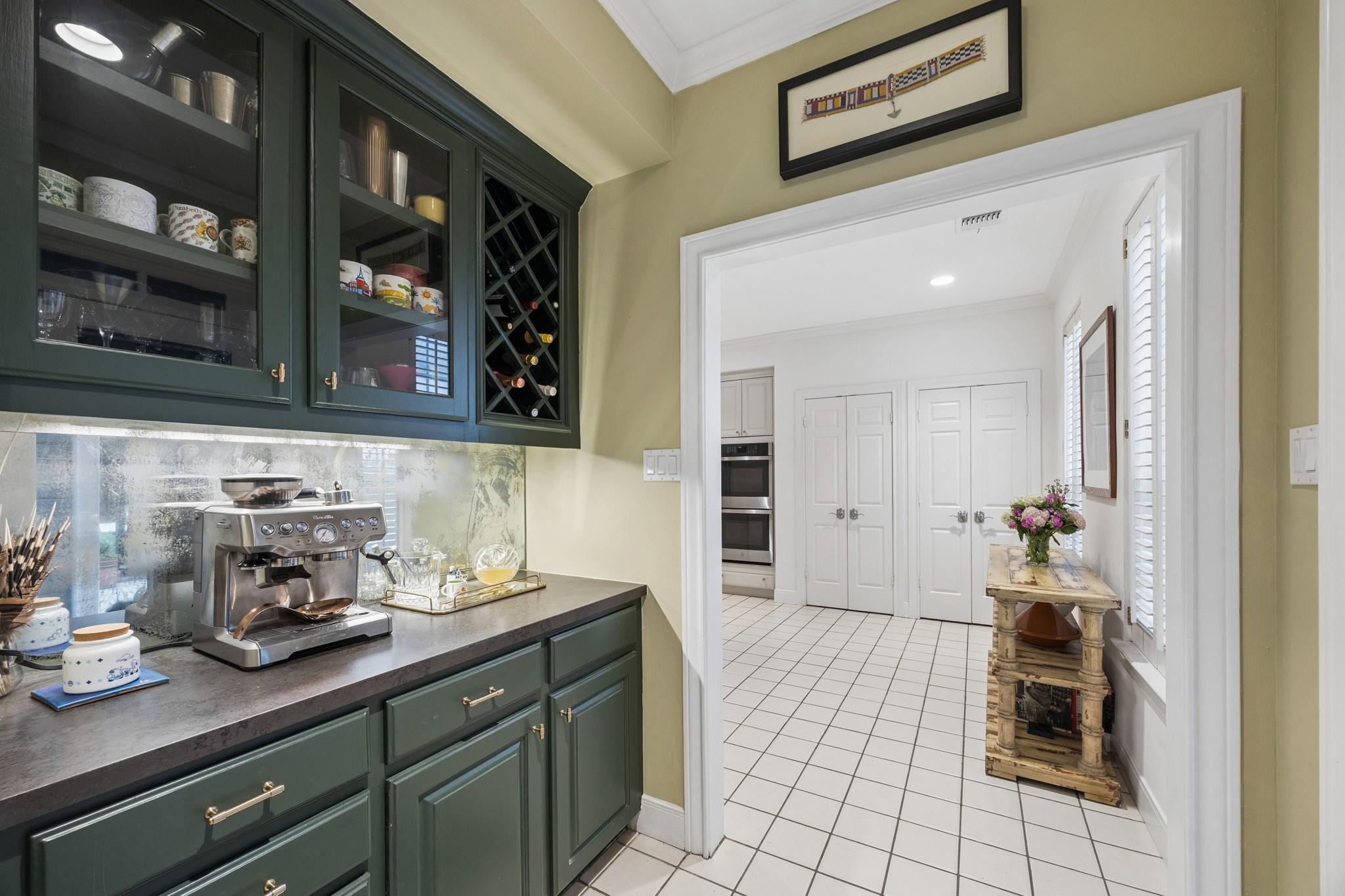 Butler’s pantry with dark green cabinets, wine rack, and espresso machine beside a bright tiled kitchen.