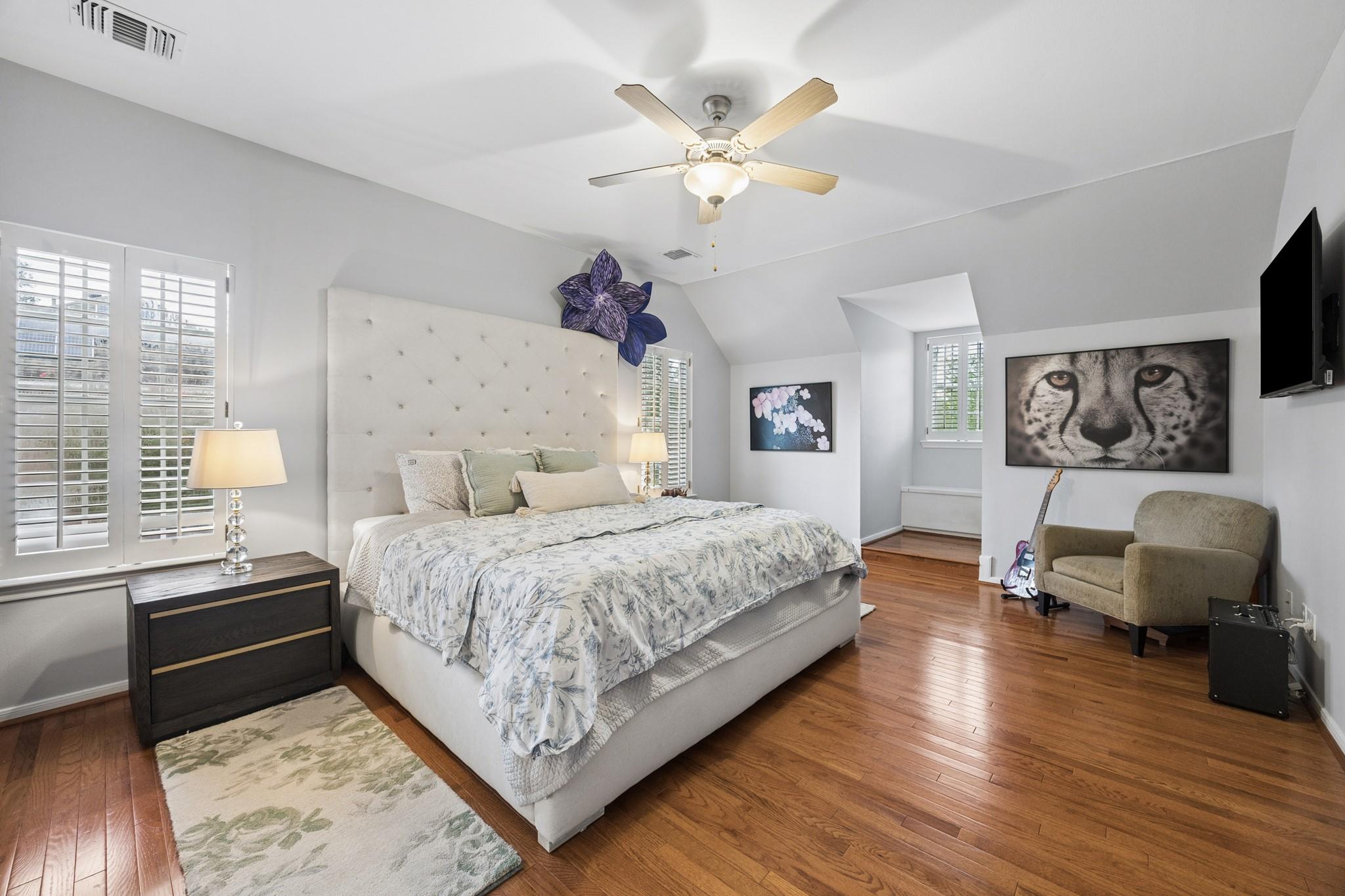 Bright bedroom with a large upholstered bed, hardwood floors, shuttered windows, and a chair beside a guitar.