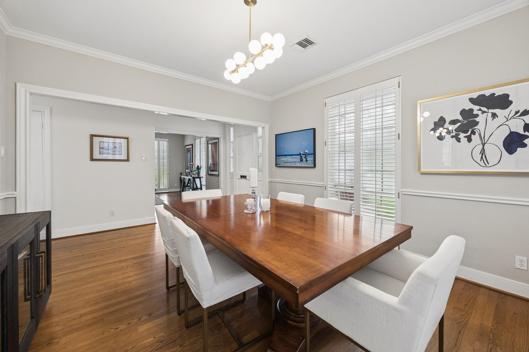Dining room with a long wood table, white chairs, plantation shutters, and a brass chandelier.