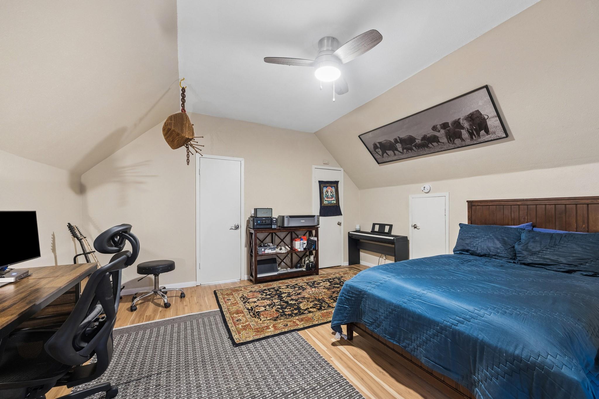 Vaulted bedroom with a blue-covered bed, wood floors, a desk, exercise bike, and digital piano.