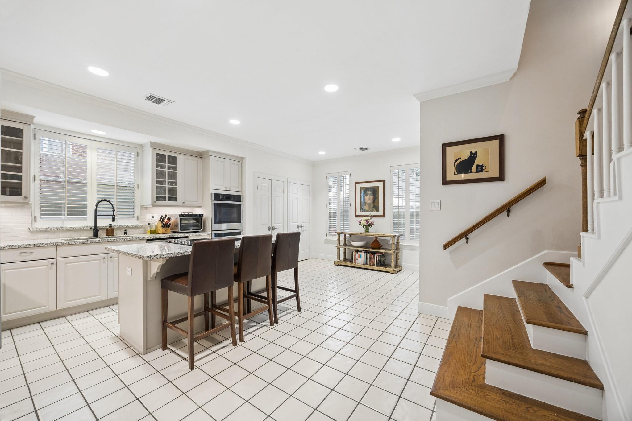 Bright white kitchen with granite island, three bar stools, tiled floor, and staircase with wooden treads.