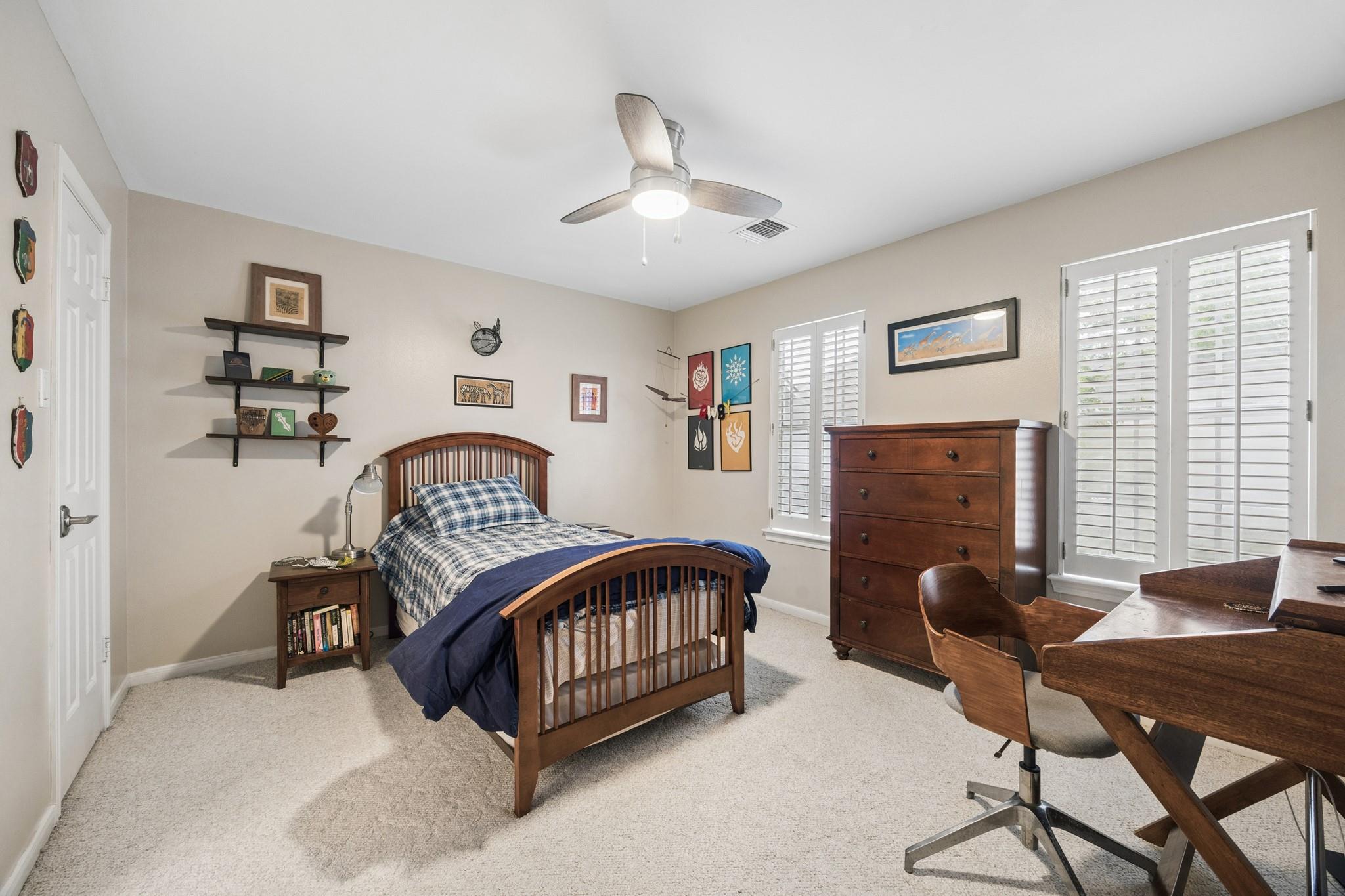 Furnished bedroom with a wooden bed, desk, dresser, and bright plantation-shuttered windows.