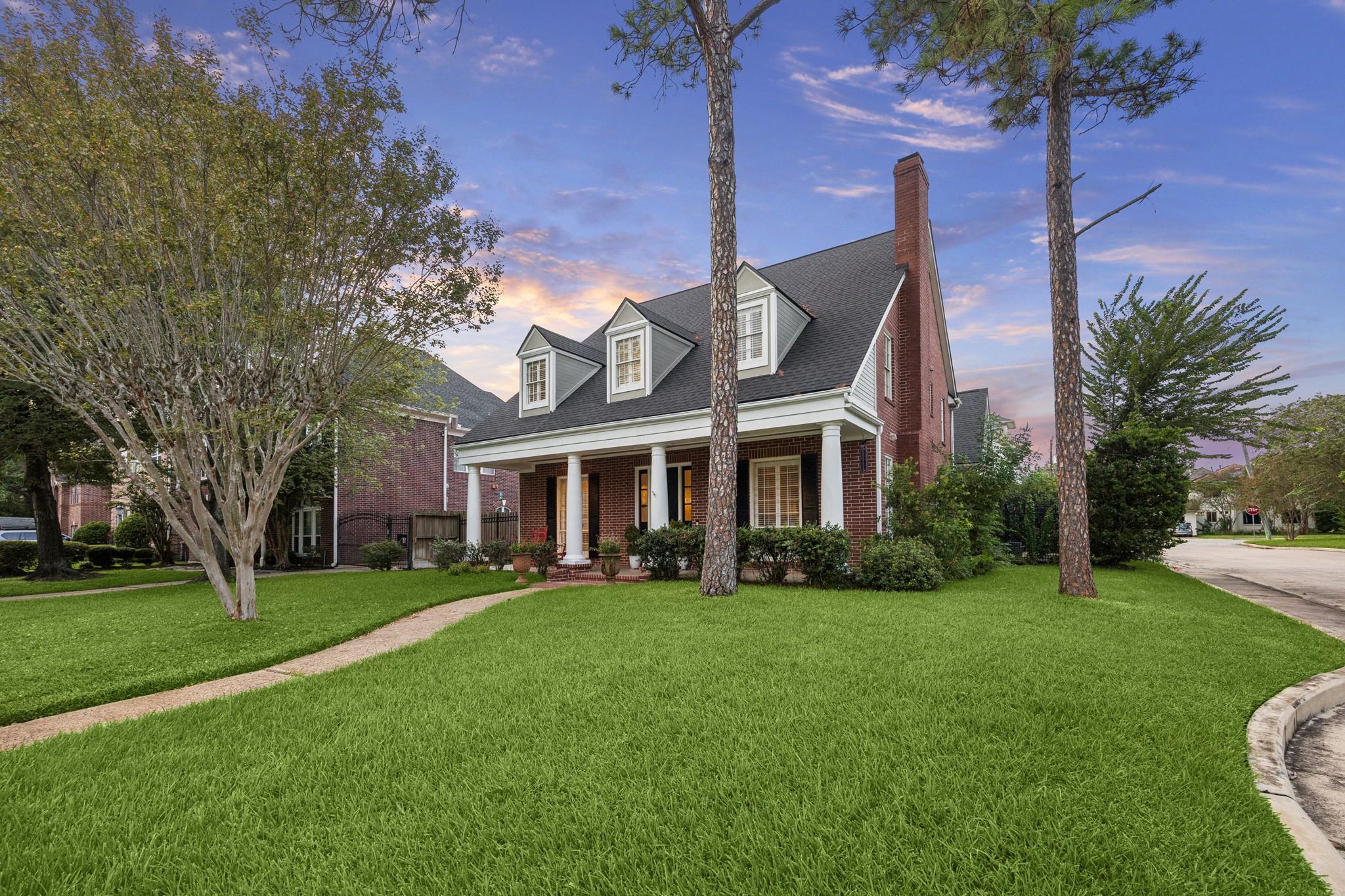 Red-brick two-story home with white porch columns, dormer windows, and a broad lawn at sunset.