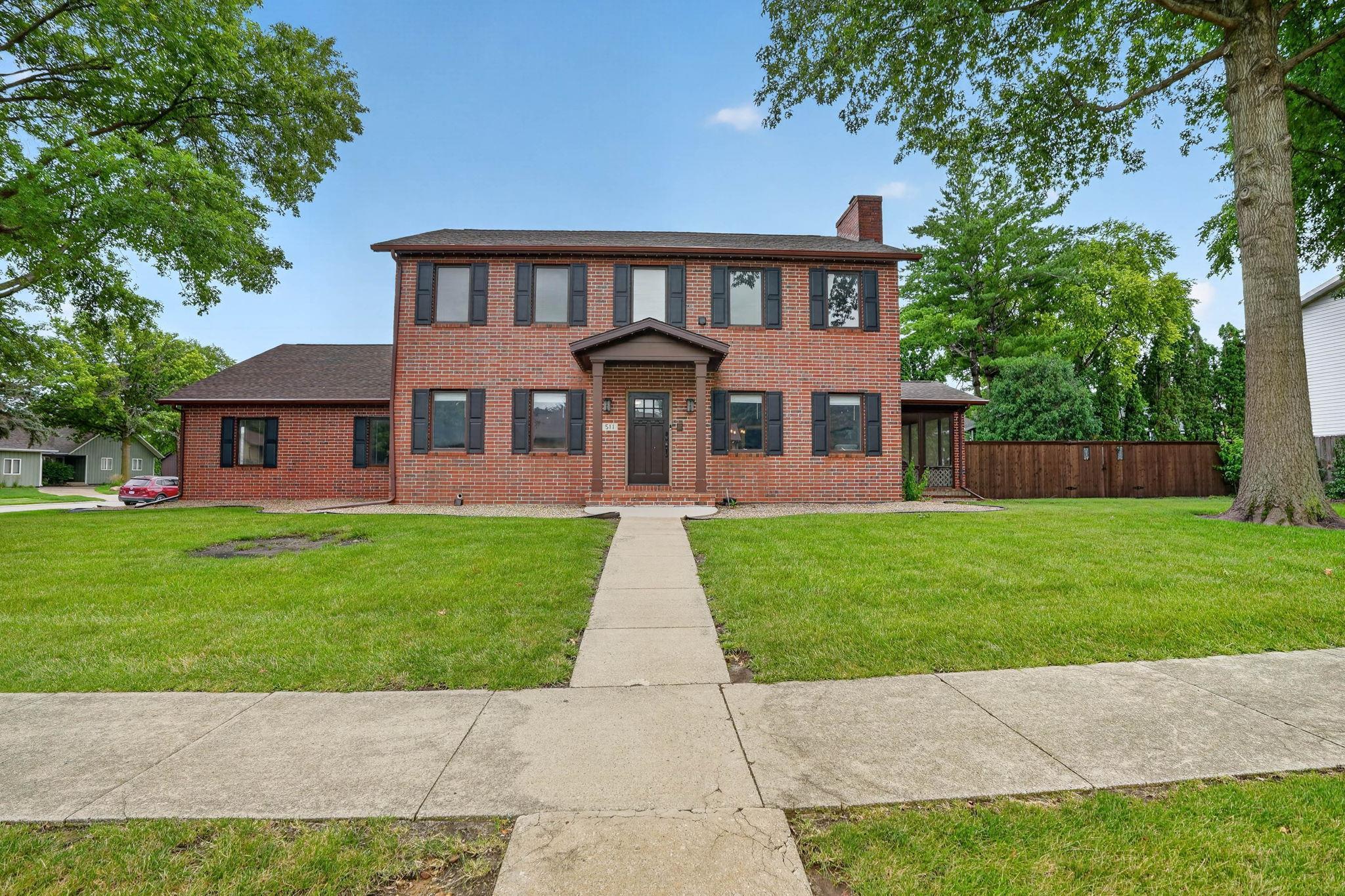 Two-story red-brick house with dark shutters, a broad lawn, and a walkway to the front door.