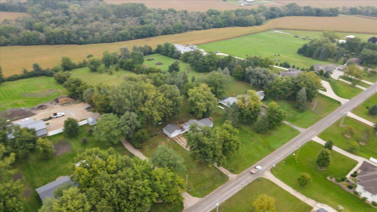 Aerial view of a rural home among mature trees, with outbuildings, open fields, and a road nearby.