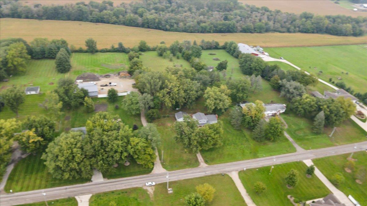 Aerial view of homes and outbuildings among mature trees beside a road and open fields.