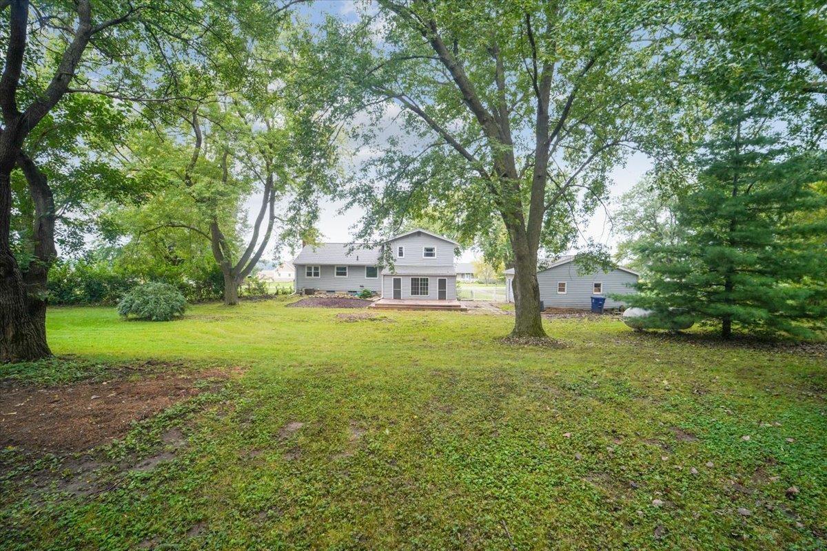 Gray two-story house and detached garage beyond a spacious lawn framed by mature trees.