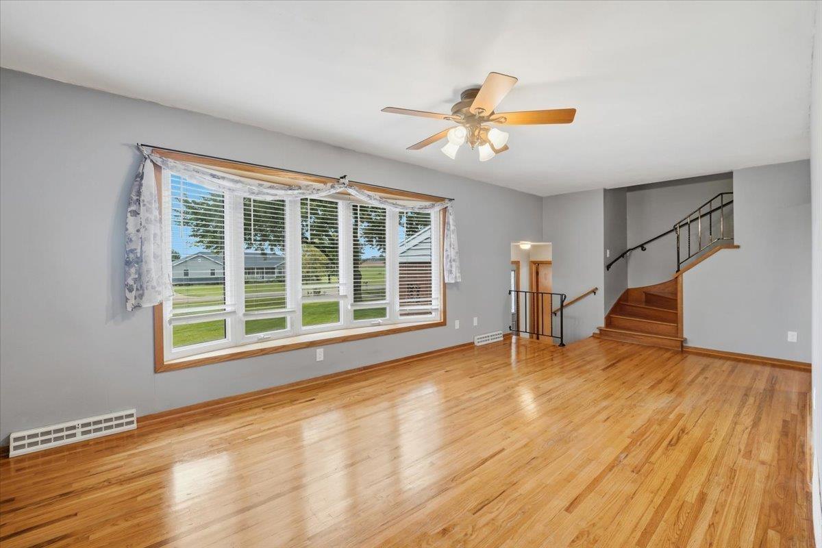 Vacant living room with polished hardwood floors, a wide window, and a wooden staircase.
