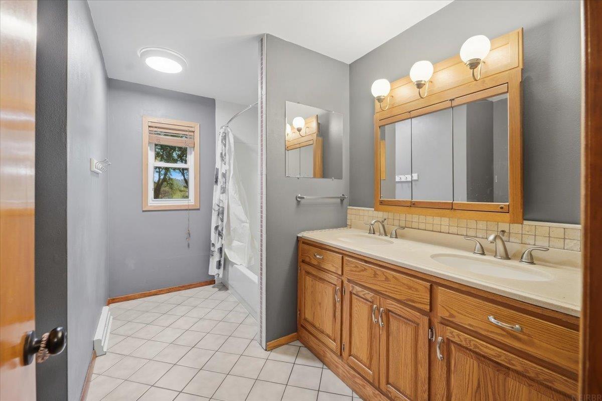 Bathroom with a double-sink oak vanity and tiled shower beside a window.