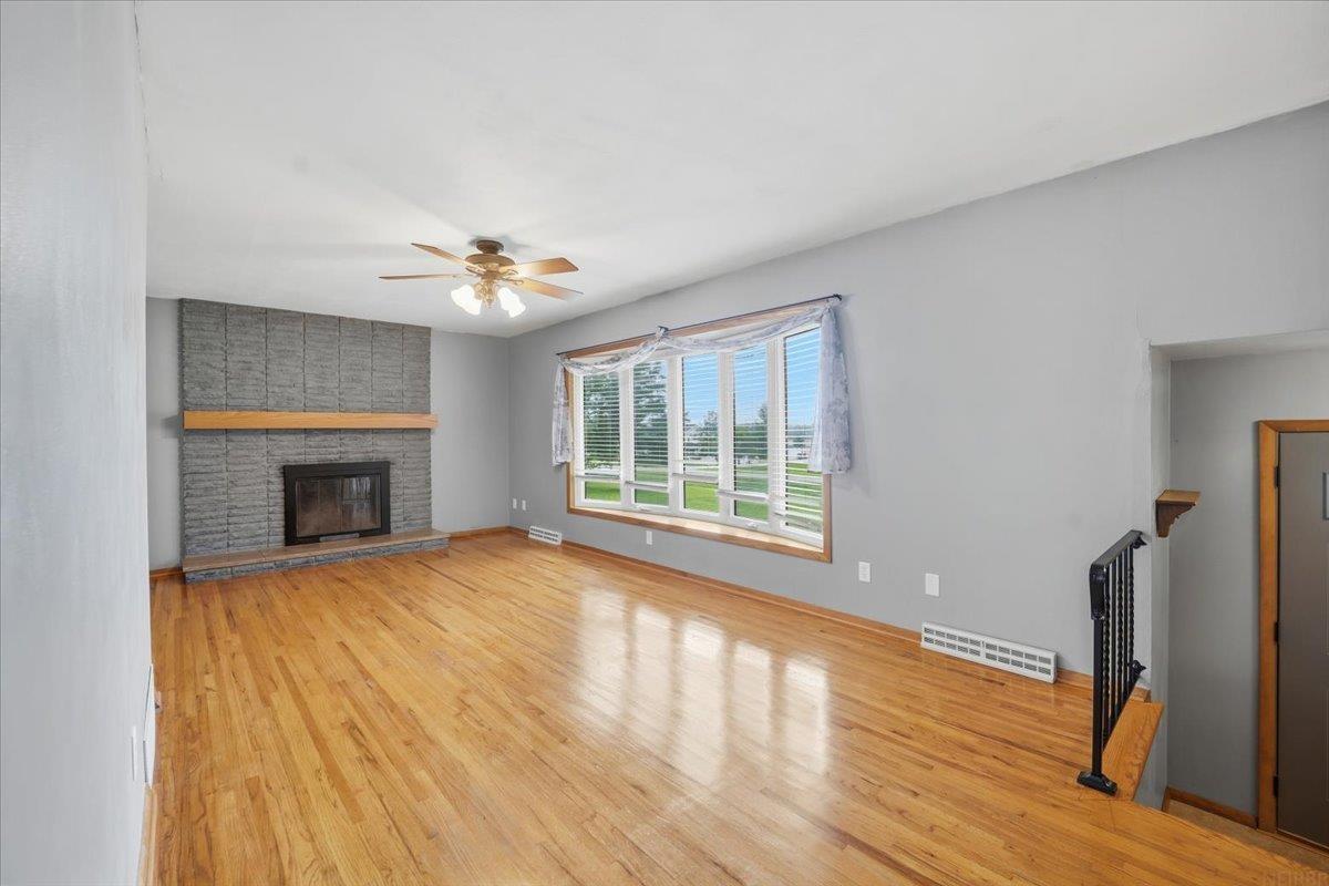 Vacant living room with hardwood floors, a brick fireplace, and a wide bank of windows.