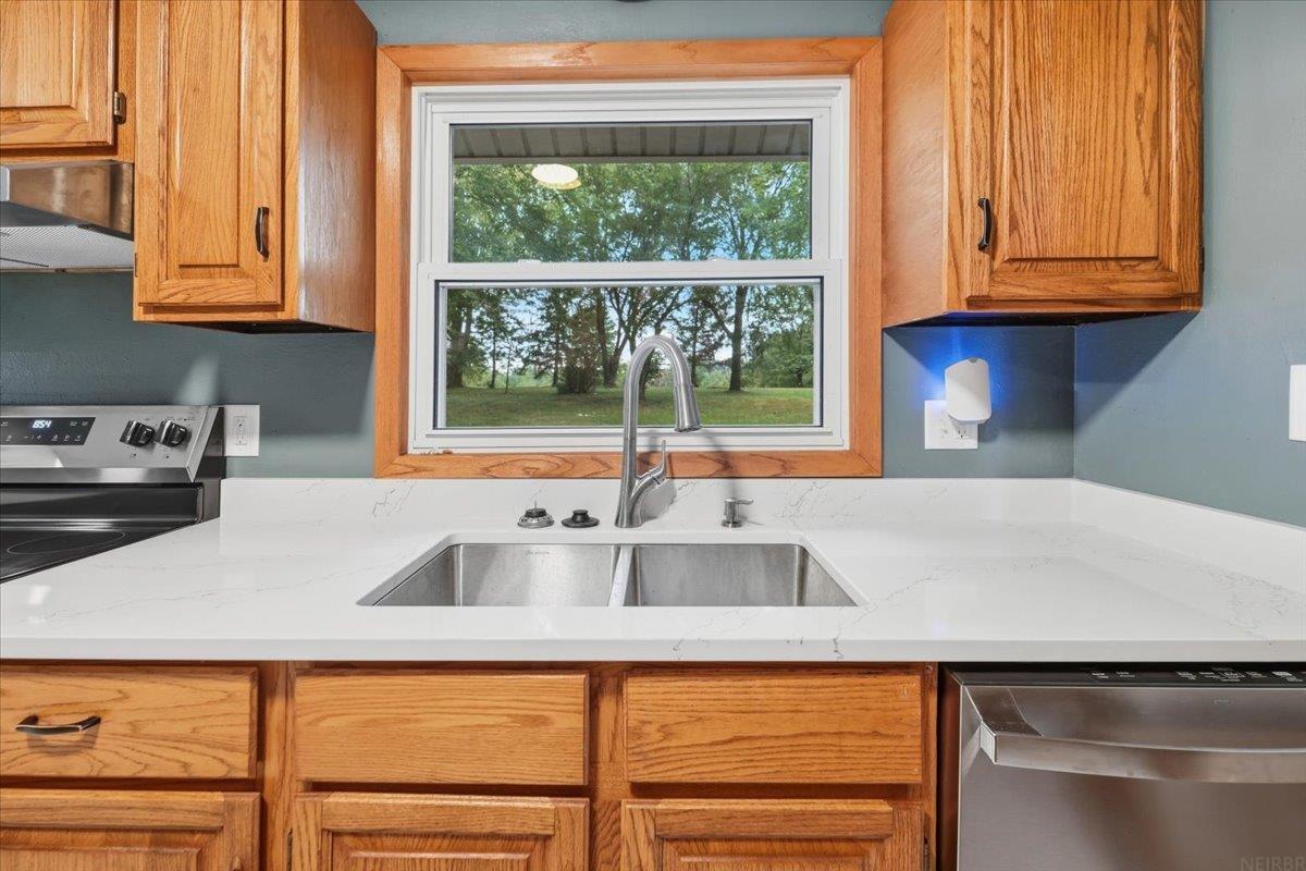 Kitchen sink beneath a window overlooking trees, with quartz counters and oak cabinets.