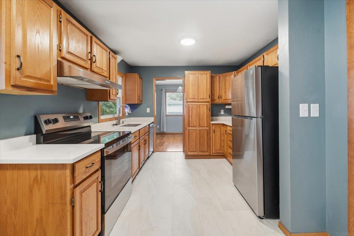 Long kitchen with wood cabinets, white counters, stainless refrigerator, and blue-gray walls.