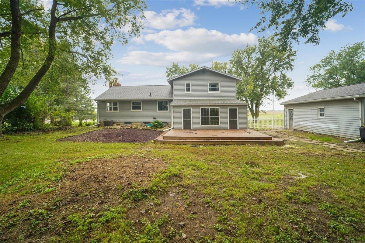 Gray two-story house with a wide wooden deck, mature trees, and a detached outbuilding in the yard.