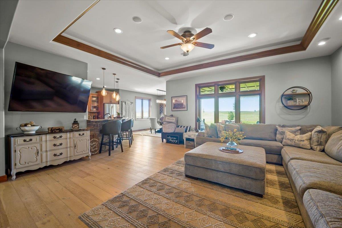 Open-plan living room with a large sectional, patterned rug, tray ceiling, and windows overlooking fields.