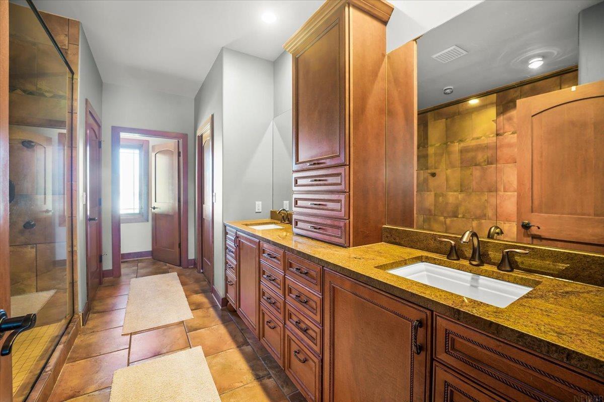 Bathroom with a long wood double vanity, stone countertop, and glass-enclosed tiled shower.