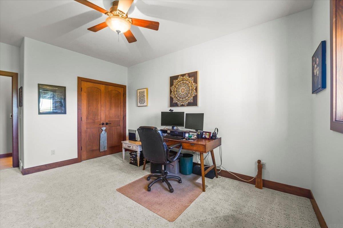 Home office with a wooden desk and monitors, black swivel chair, ceiling fan, and wooden double doors.