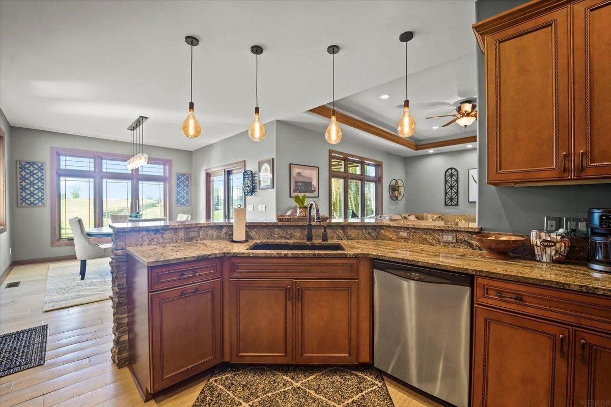 Kitchen with wood cabinetry, granite counters, stainless dishwasher, and pendant lights beside large windows.