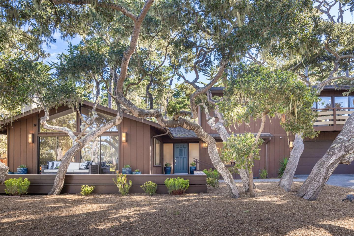 Brown home with a blue front door and deck beneath mature twisting trees.