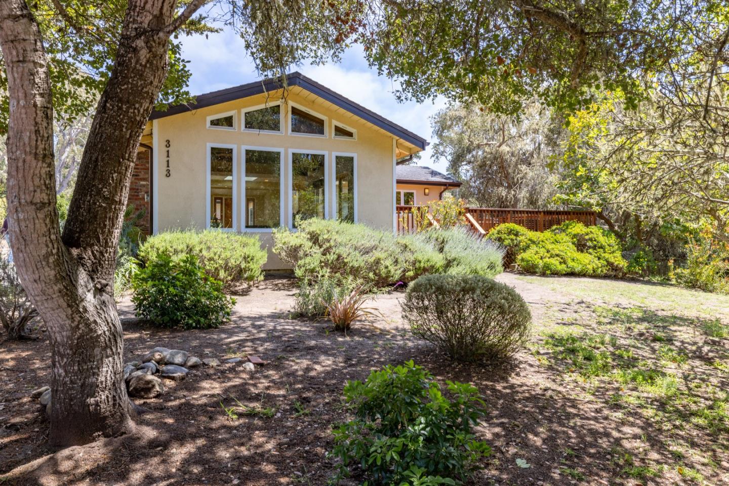 Home with tall front windows and a wooden deck, surrounded by mature trees and shrubs.