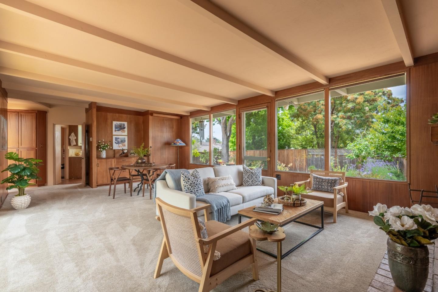 Living room with wood-paneled walls, exposed beams, and large windows overlooking a leafy garden.