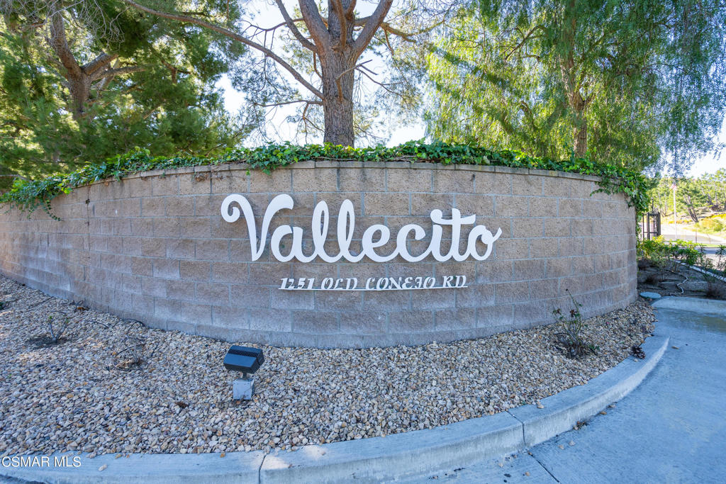 Vallecito entrance sign on a curved stone wall beneath mature trees.