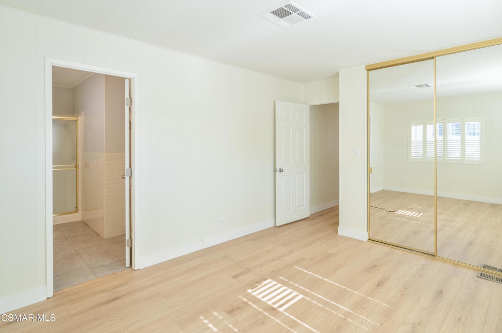 Empty bedroom with mirrored closet doors, light wood flooring, and an adjoining bathroom.