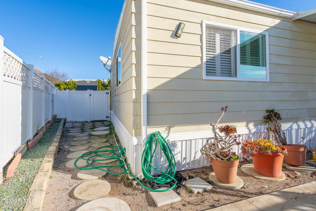Side yard beside a beige home, with stepping stones, white fencing, potted plants, and coiled garden hoses.
