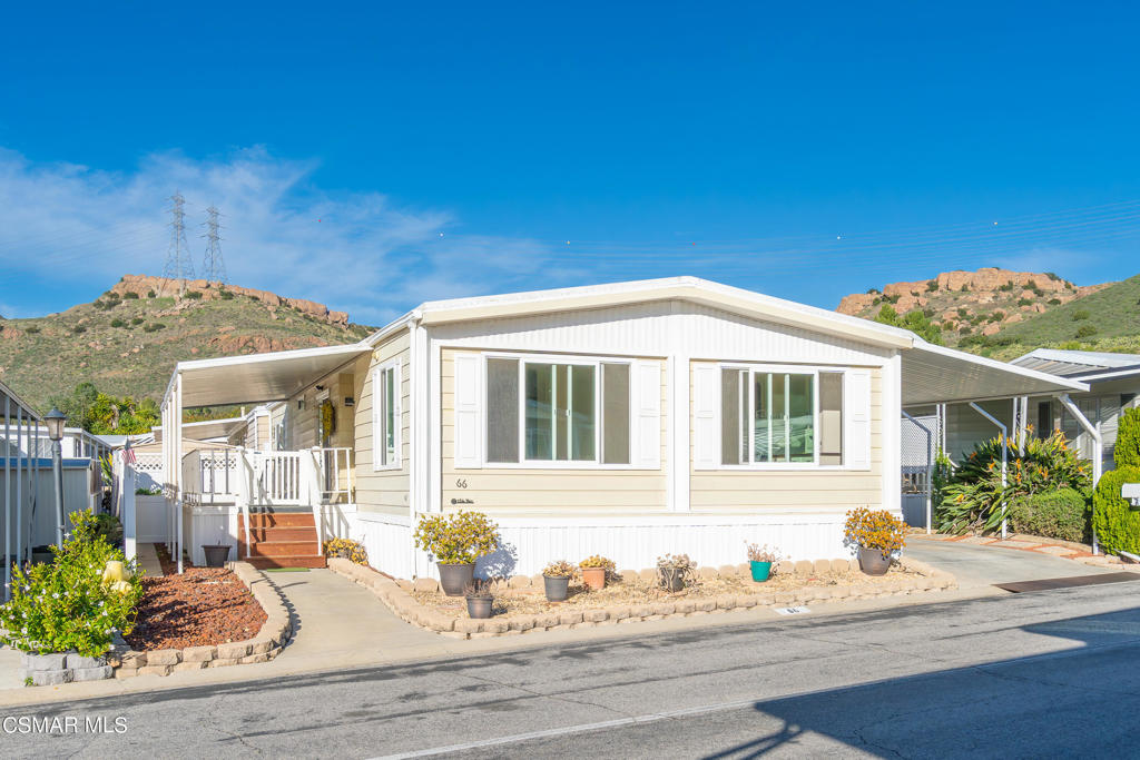 Light-colored manufactured home with covered parking, landscaped entry, and rocky hills behind it.