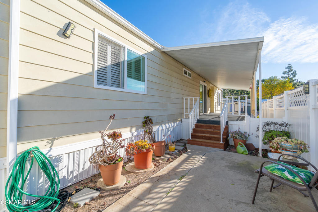 Covered side patio with steps, white fencing, potted plants, and a green garden hose.