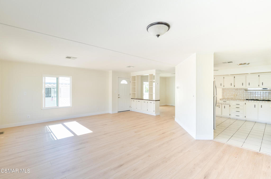 Vacant living area with pale wood floors, a large window, and an adjoining white kitchen.