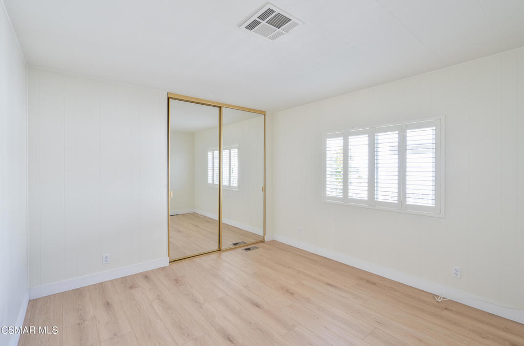 Empty bedroom with mirrored closet doors, plantation shutters, and light wood flooring.