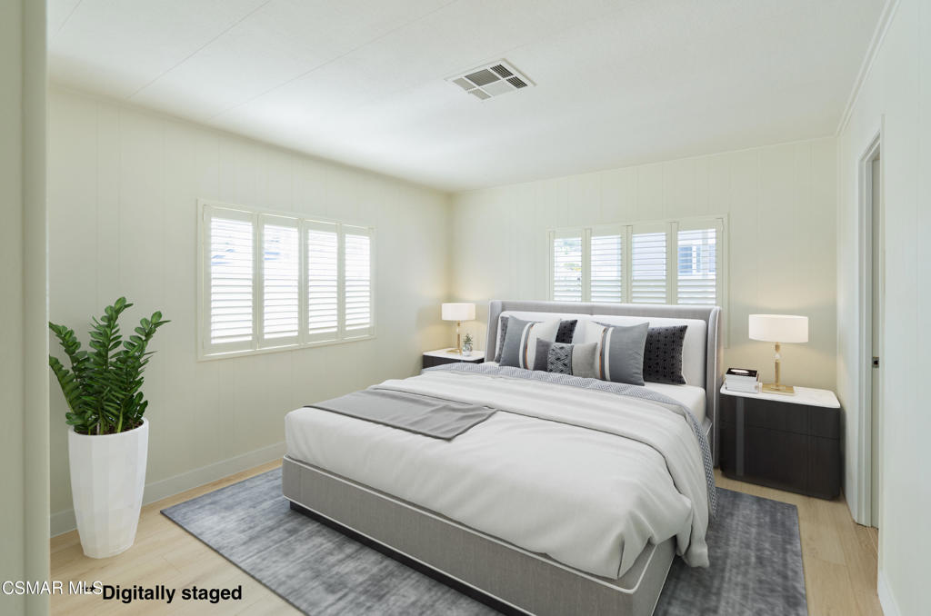 Staged bedroom with a large upholstered bed, gray rug, shuttered windows, and potted plant.