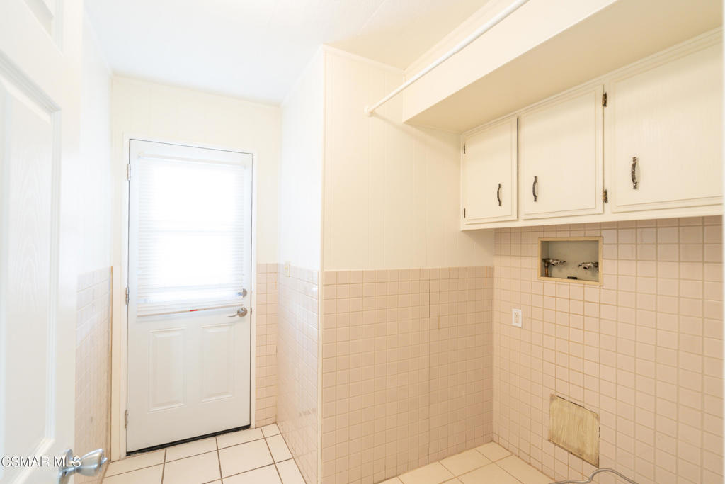 Empty laundry room with upper cabinets, washer hookups, and pale tiled walls and floor.