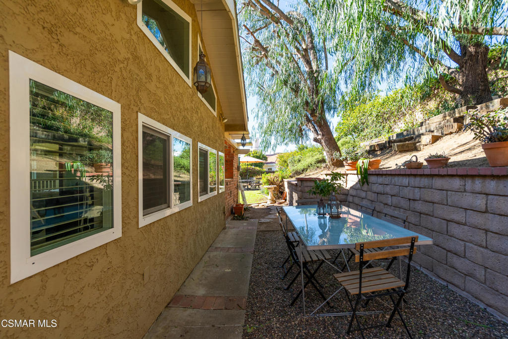 Side patio beside a tan stucco house, with a glass-top dining table and leafy trees.