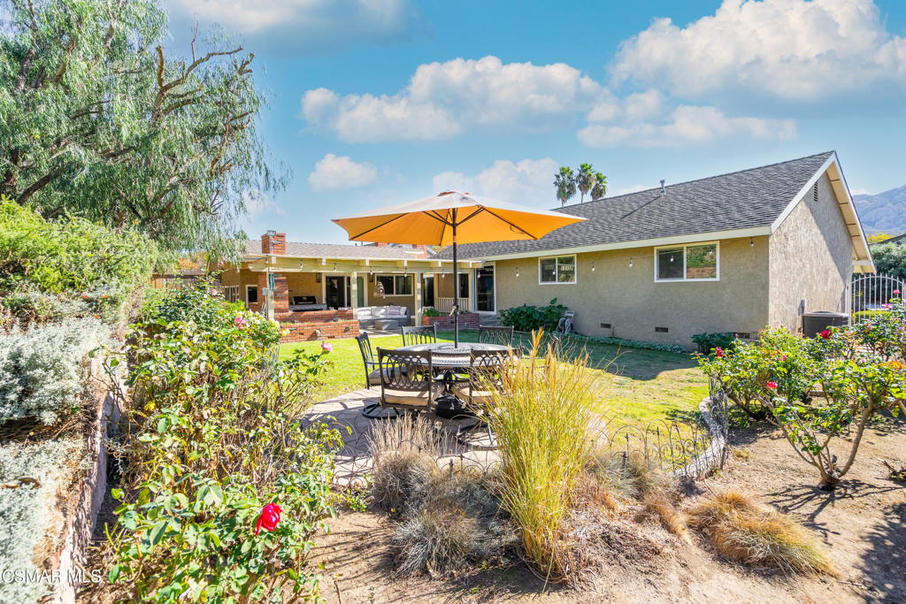 Home’s backyard with a stone patio, outdoor dining set, orange umbrella, lawn, and landscaped garden.