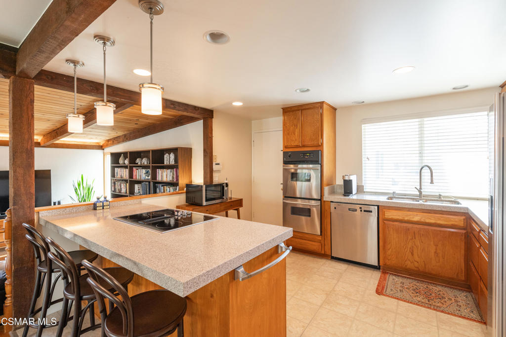 Kitchen with a large cooktop island, warm wood cabinetry, stainless steel appliances, and exposed beams.