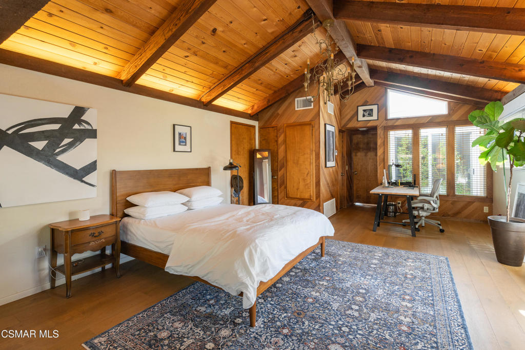 Bedroom with a vaulted wood-beam ceiling, blue patterned rug, and desk beside the windows.
