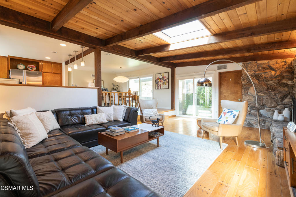 Living room with a black leather sectional, stone fireplace, and skylight beneath exposed timber beams.
