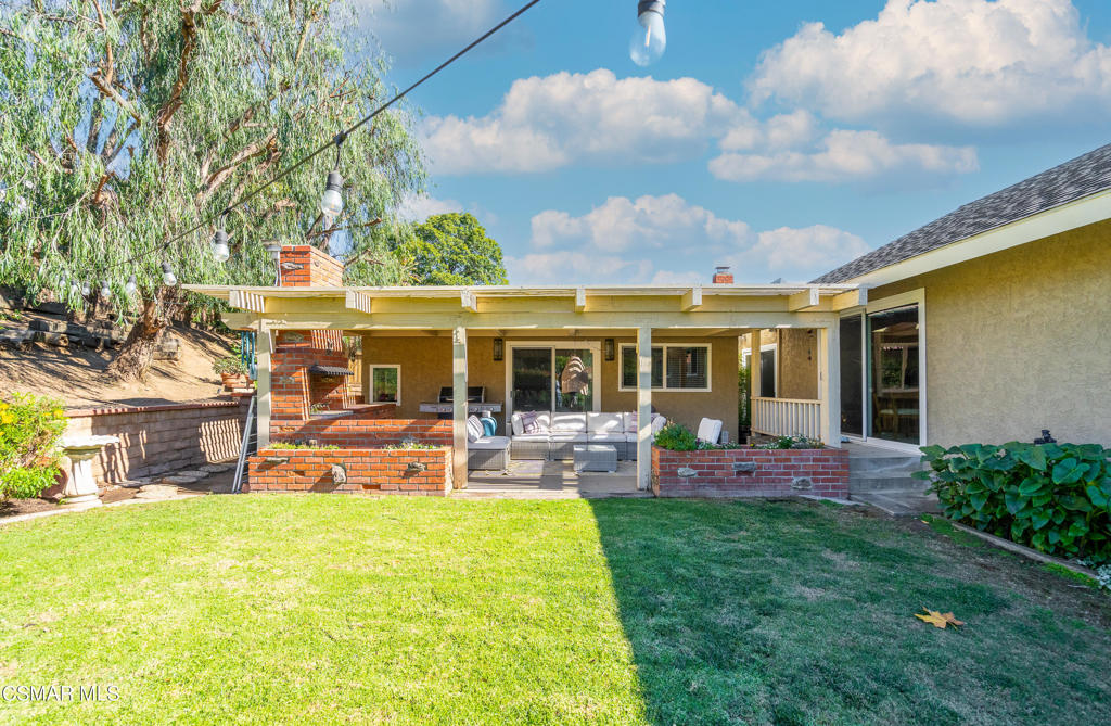 Home’s covered backyard patio with outdoor seating, brick fireplace, and green lawn.