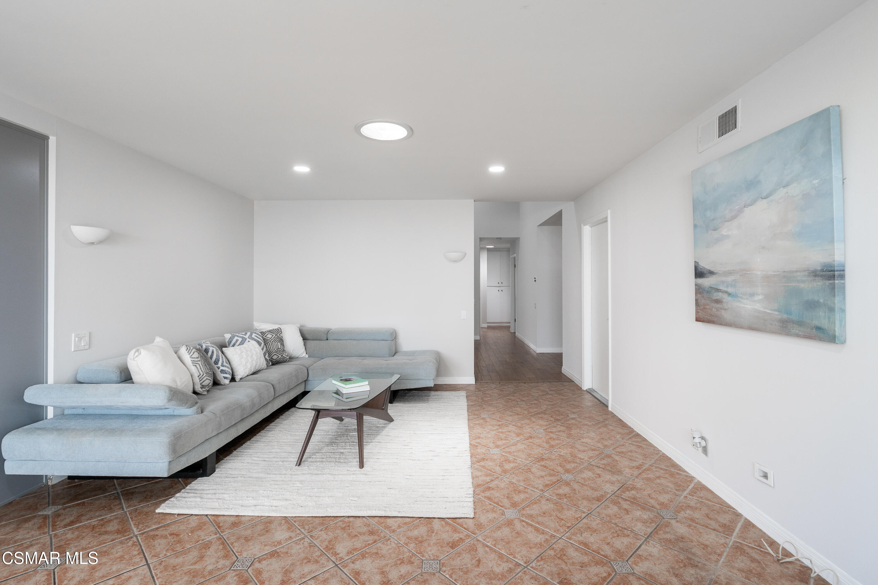 Living room with a gray sectional, white rug, terracotta tile floor, and blue-toned wall art.