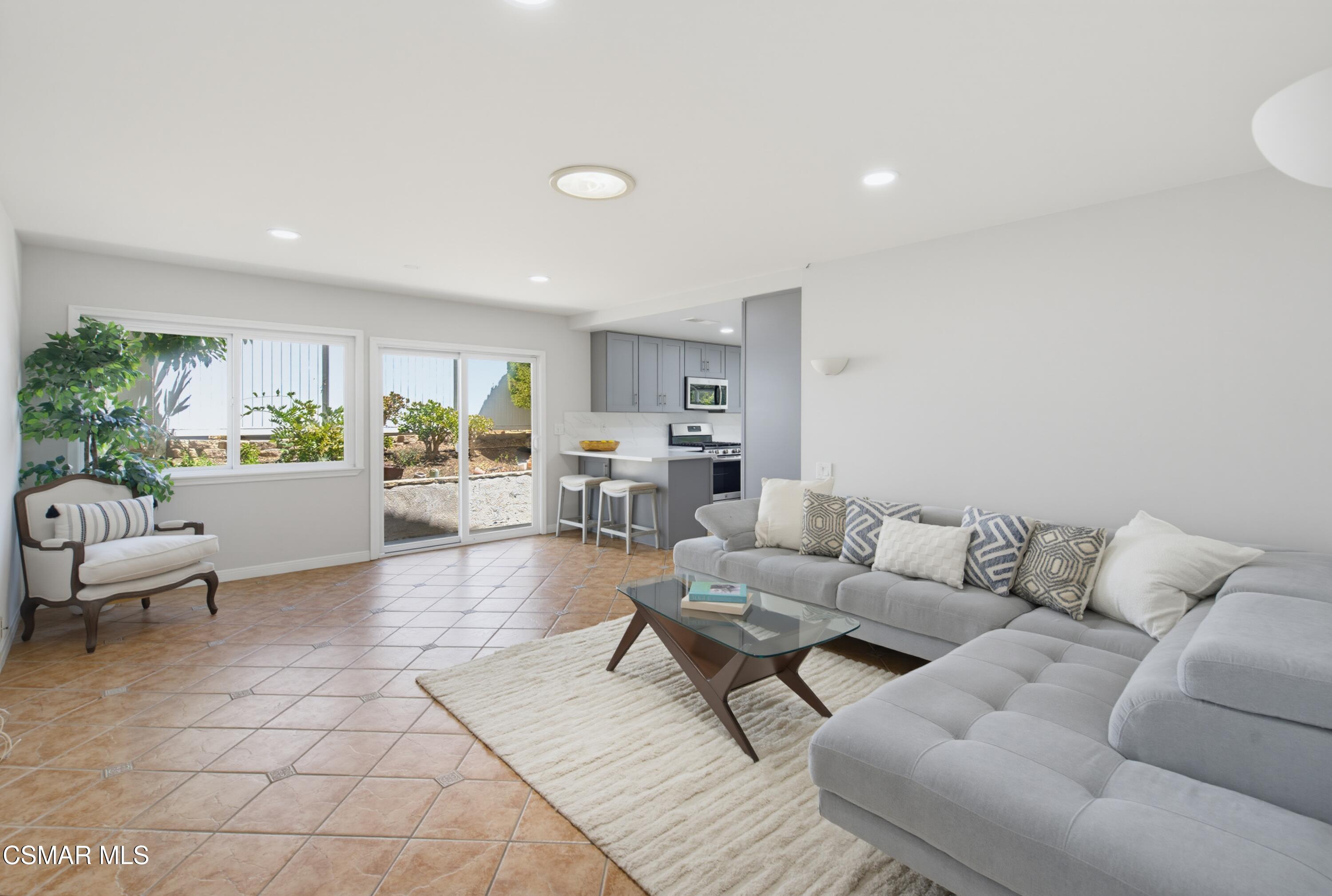 Living room with a gray sectional and glass coffee table, opening to a gray-cabinet kitchen.