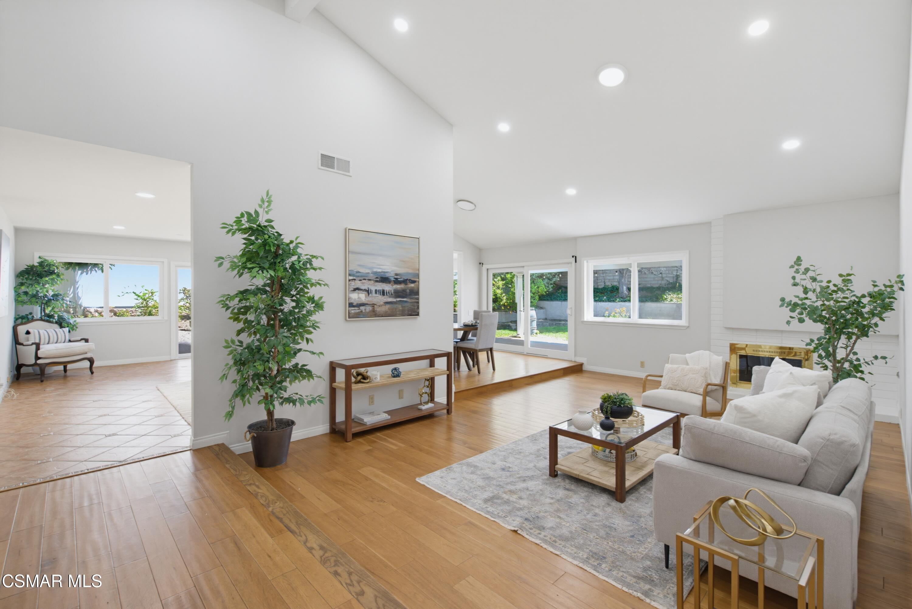 Open-plan living room with hardwood floors, white seating, a fireplace, and windows overlooking the garden.