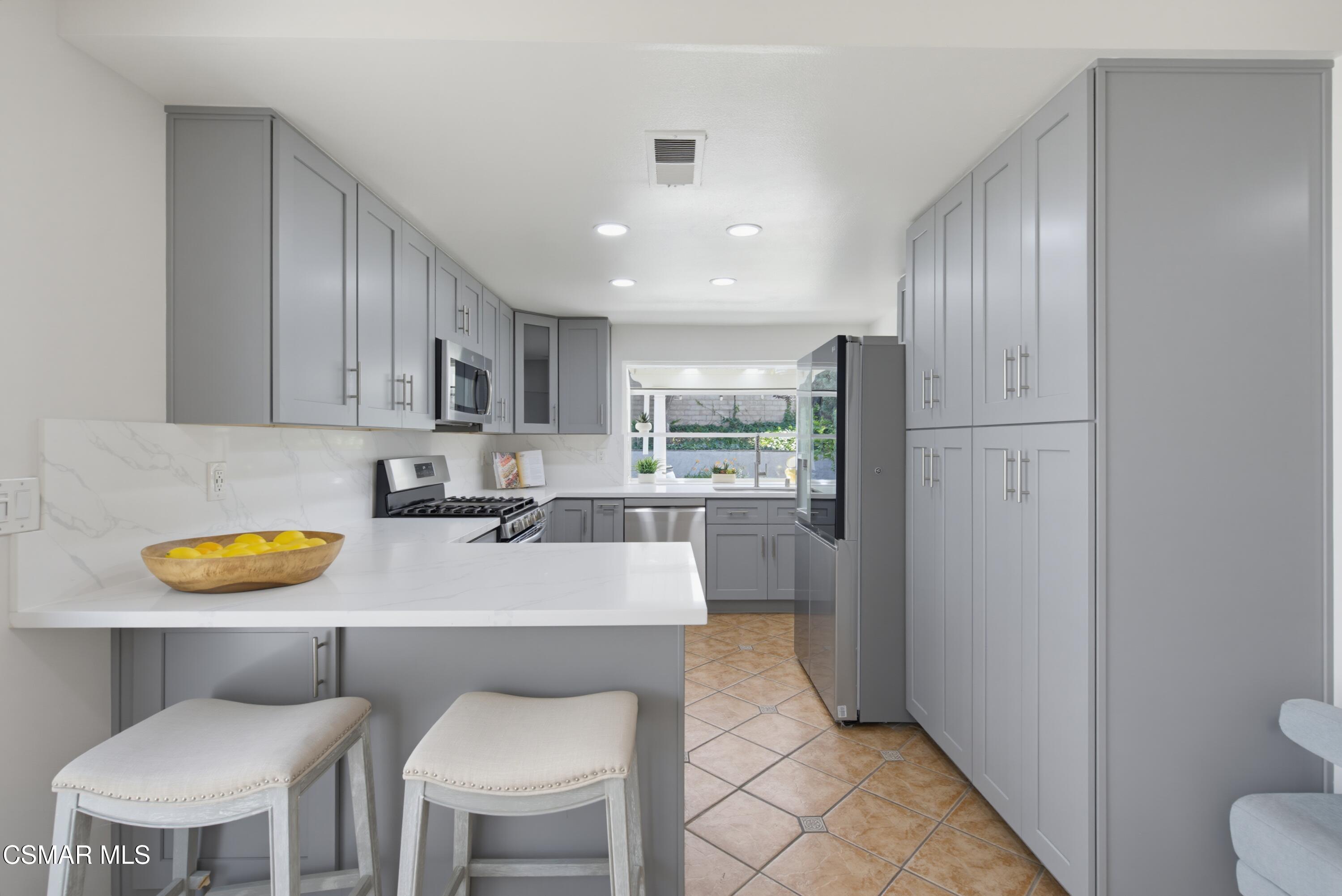 Kitchen with gray cabinets, a white peninsula, two upholstered stools, and a window overlooking a patio.