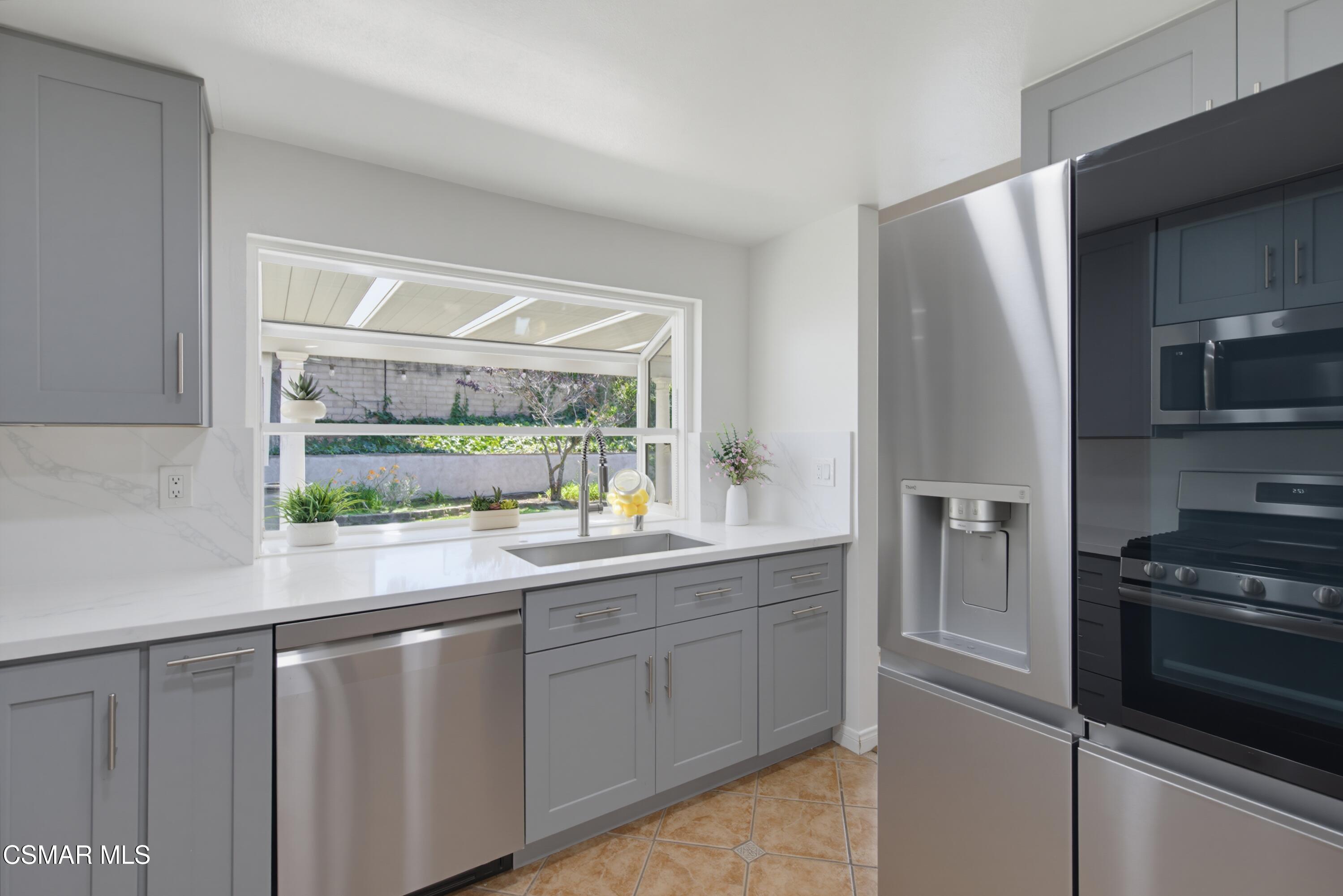 Bright kitchen with gray cabinets, stainless steel appliances, and a sink beneath a garden-facing window.