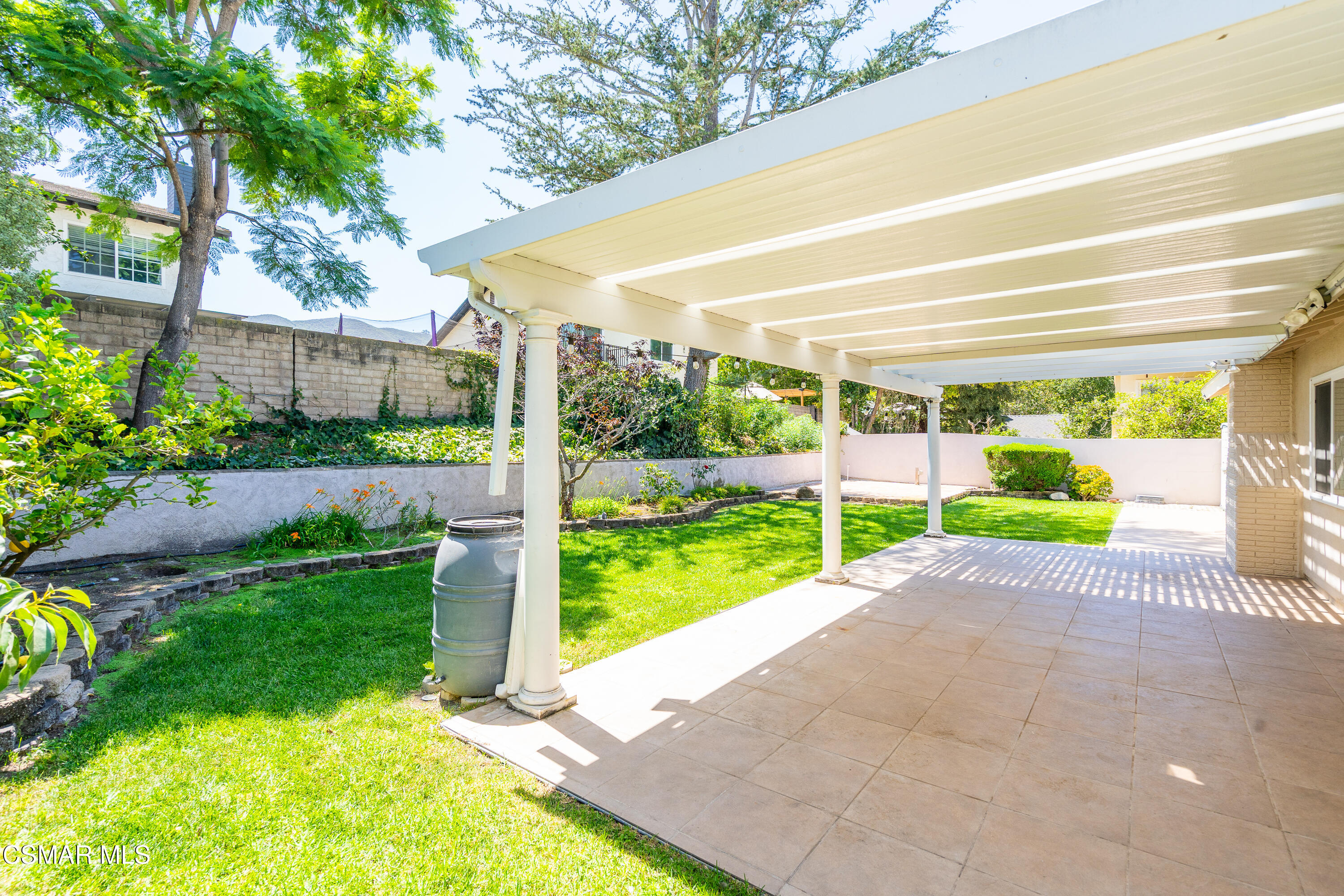 Covered pergola patio beside a green lawn, with tiled flooring and landscaped garden beds.