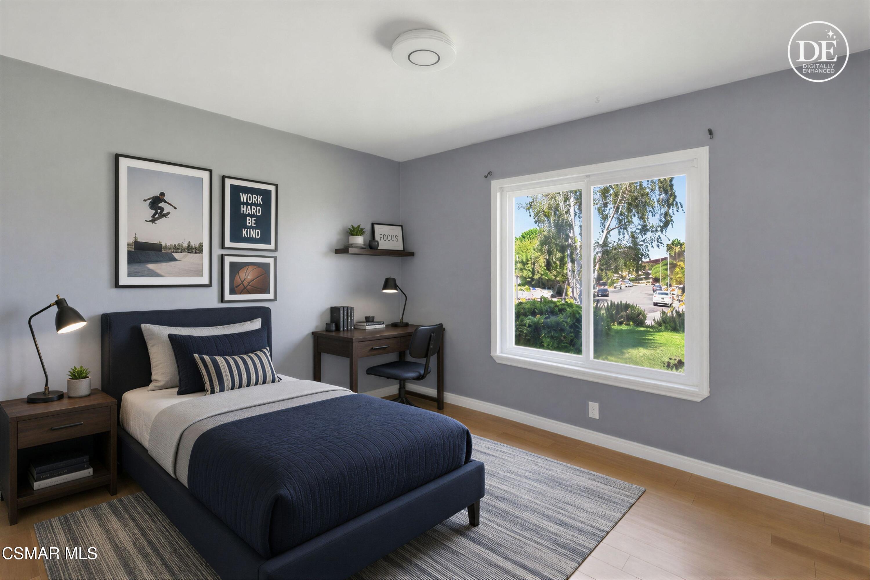 Staged bedroom with a navy bed, desk, and large window overlooking trees and a residential street.