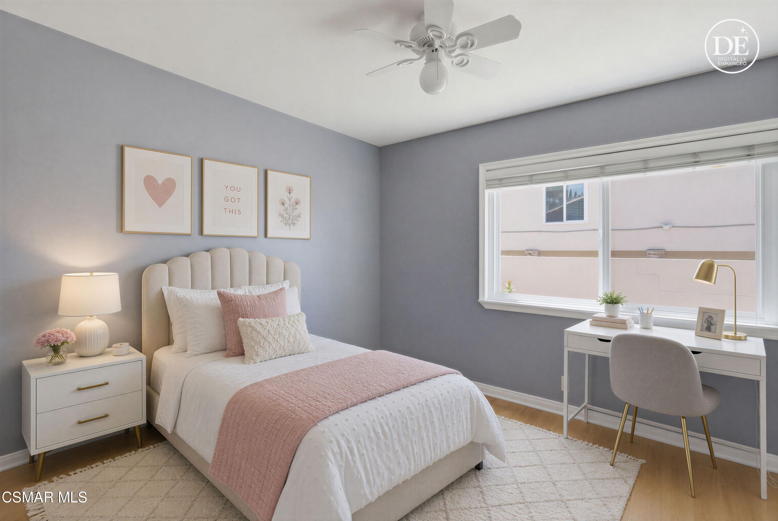 Staged bedroom with blush bedding, gray walls, and a white desk beneath a large window.